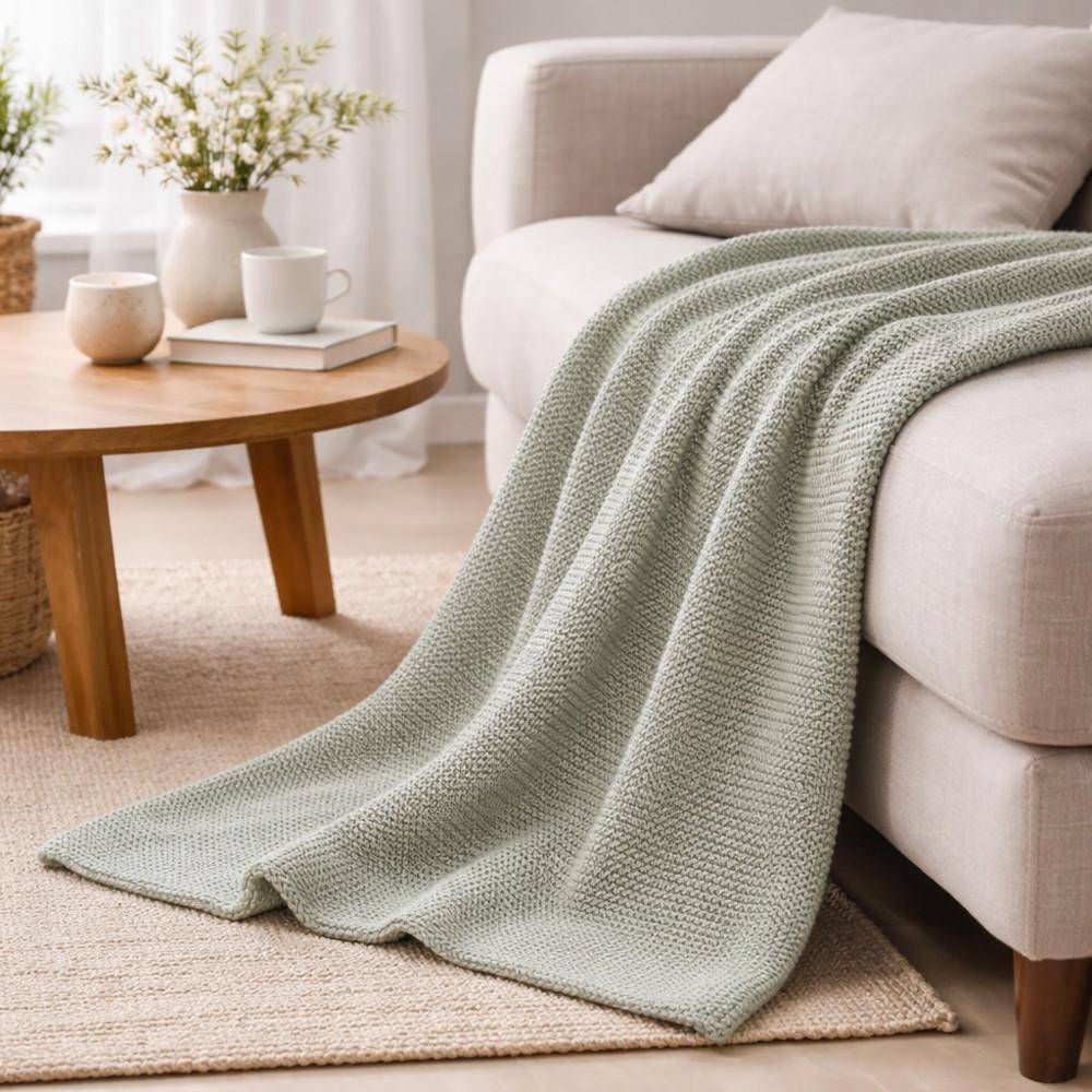 Grünes Strickplaid in Garden Green auf Sofa drapiert in hellem Wohnzimmer mit Holztisch und dekorativen Accessoires